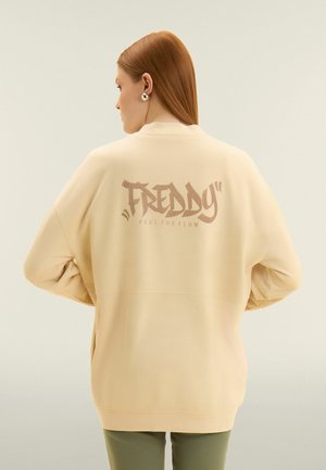 Mujer con cabello rojo liso que lleva una sudadera beige con el texto "FREDDY FEEL THE FLOW" en la espalda, combinada con pantalones verde oliva.