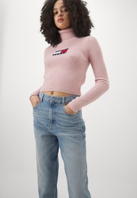 Roza rebrasta turtleneck pulover z skrajšanim dizajnom in logotipom. V kombinaciji z svetlo modrimi hlačami z visokim pasom.