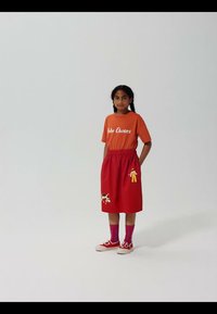 Pige med fletninger står iført en orange "Bobo Choses" t-shirt, rød nederdel med legende lapper, lilla sokker og røde sneakers.