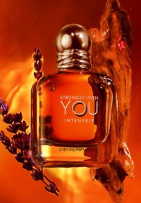 Parfymeflaske merket "Stronger With You Intensely" av Emporio Armani, omgitt av lavendelkvaster og et trestykke på en oransje bakgrunn.