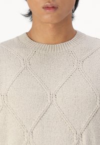 Gros plan sur une personne portant un pull tricoté beige clair avec un motif en losange et un col rond côtelé.