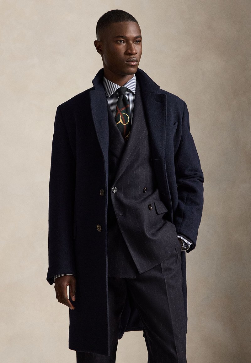Homme portant un costume sombre croisé, un pardessus bleu marine, une chemise rayée et une cravate à motifs, debout avec une main dans la poche sur un fond uni.