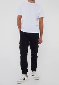 Bílé bavlněné tričko s kulatým výstřihem doplněné tmavými kalhotami typu cargo s kapsami po stranách a elastickými manžetami, doplněné bílými teniskami.