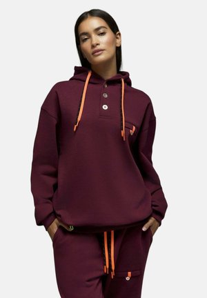 Femme portant un sweat à capuche grenat et un pantalon de survêtement avec des cordons orange et des boutons, mains dans les poches, debout devant un fond clair.