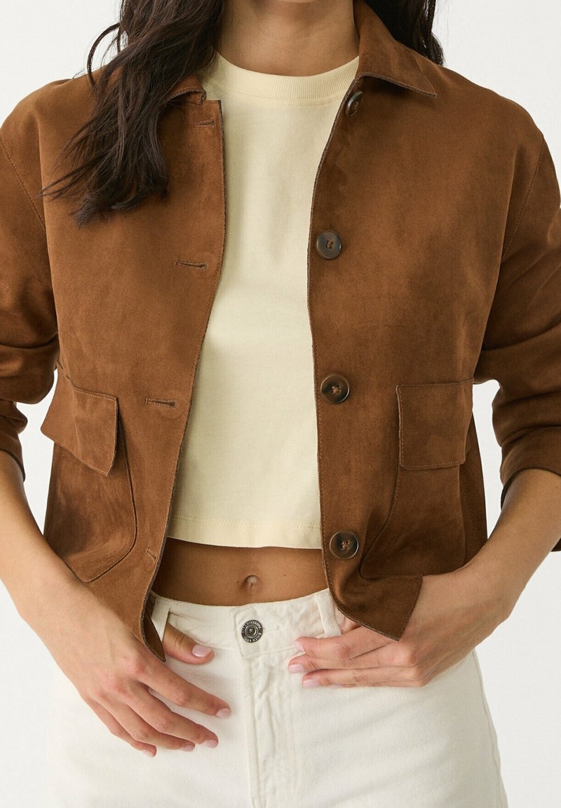 Woman wearing a brown suede button-up jacket over a cream crop top paired with high-waisted white pants, hands near waist.