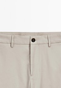 Beige bomullbyxor med flat front, featuring en enkel knappstängning och bälteshällor. Slät textur med subtila sömdetaljer.