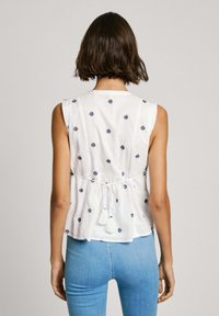 Haut sans manches blanc avec des motifs brodés marine, détail de lien ajustable à l'arrière et coupe décontractée, associé à un jean bleu clair.