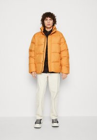 Oranje pufferjack met een boxy ontwerp, hoge kraag en drukknoopsluiting, gecombineerd met een donker geruite shirt en lichtgekleurde broeken.