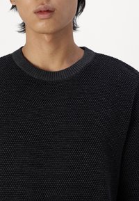 Maglione nero lavorato a maglia con una superficie texturizzata, scollatura tonda e polsini a costine. Il materiale sembra morbido e spesso, adatto per il clima più fresco.