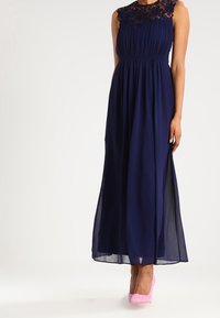 Navy maxi dress with lace neckline and pleated skirt. Sheer fabric with a side slit, paired with pink high heels.