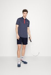 Polo rayé marine avec des accents de boutons rouges, associé à un short marine. Le modèle tient une raquette de tennis et porte des baskets marines et blanches.