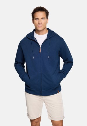 Hombre con cabello corto y castaño, vistiendo una sudadera con capucha azul marino y cremallera sobre una camiseta blanca y pantalones cortos beige, de pie con las manos en los bolsillos.