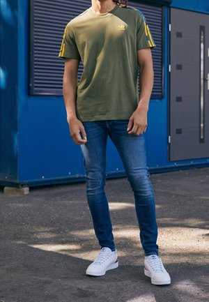 Vihreä puuvillainen t-paita, jossa on keltainen Adidas-logo ja raidalliset hihat, yhdistetty sinisiin farkkuihin ja valkoisiin lenkkareihin, seisomassa teksturoidulla pinnalla.