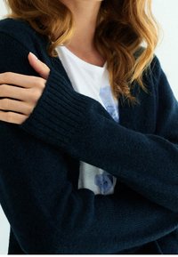 Mörkblå stickad kofta med ribbade muddar, bärs över en ljus skjorta med ett blått mönster. Mjuk textur, ärmar i midjelängd.