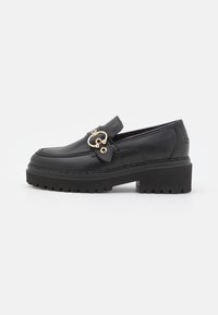 Mocasines de cuero negro con una suela gruesa negra, que presentan una hebilla decorativa dorada y un detalle de costura a lo largo de los bordes.