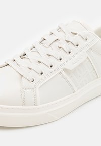 Bílé kožené tenisky s texturovanými bočními panely, tonální tkaničky, embosované logo. Vyznačují se nízkoprofilovou gumovou podrážkou a minimalistickým designem.