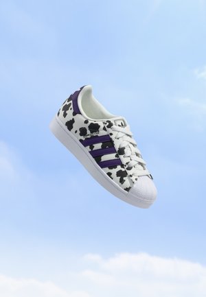 Baskets blanches avec imprimé vache noir, bandes violettes et lacets blancs flottant sur un fond de ciel bleu.