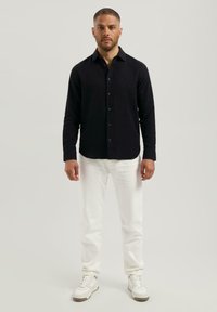Man die rechtop staat, gekleed in een zwart overhemd met knopen, witte broek en witte sneakers tegen een licht achtergrond.