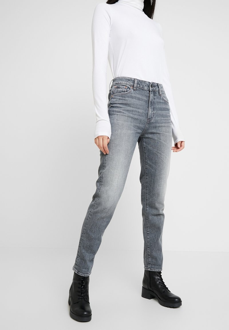 Personne portant un col roulé blanc à manches longues, un jean gris taille haute et des bottines noires à lacets, debout sur un fond uni.