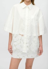 Robe blanche à manches courtes, à boutons sur le devant, avec un ourlet en dentelle ornée de motifs floraux. Poche sur le côté gauche de la poitrine.