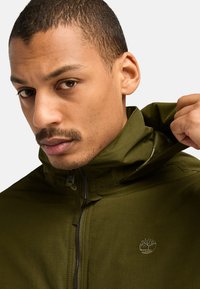 Olivgrüne wasserdichte Jacke mit hohem Kragen, Reißverschluss und gesticktem Logo auf der Brust. Glatte Textur und leichtes Design.