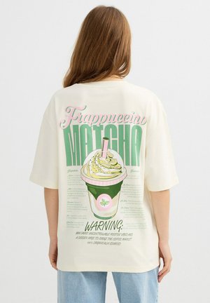 Persona con pelo largo que lleva una camisa blanca con un gráfico colorido de frappuccino de matcha y texto en la espalda.