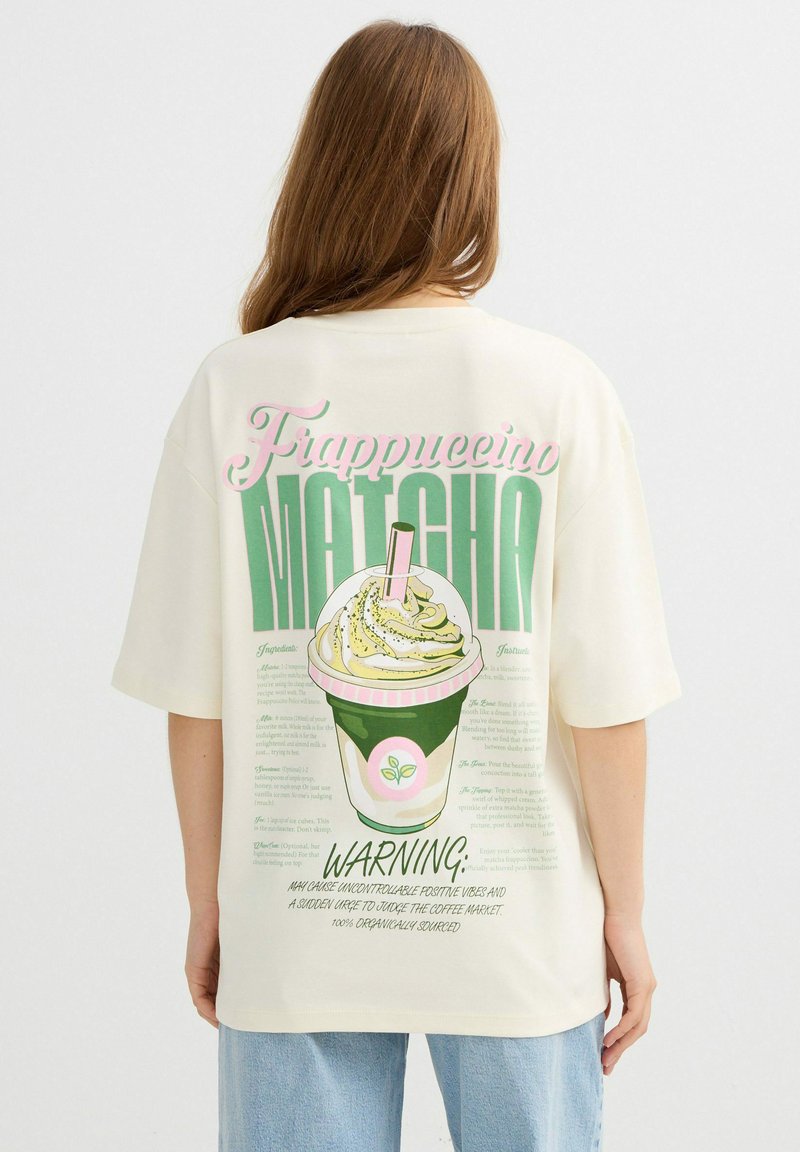 Persona con pelo largo que lleva una camisa blanca con un gráfico colorido de frappuccino de matcha y texto en la espalda.