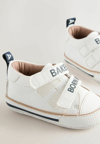 Biele tenisky pre batoľatá so suchými zipsami označené "BAKER" a "BORN." Mäkký materiál, zaoblený tvar a štruktúrovaná gumová podrážka s námorníckym akcentom.
