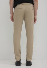 Pantaloni beige con vestibilità sartoriale, dotati di due tasche posteriori e dettagli di cucitura discreti. Abbinati a scarpe nere, senza motivi visibili.
