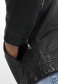 Hand in de zijzak van een zwart leren jack met een zichtbare zilveren rits en blauwe spijkerbroek eronder.