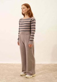 Pruhovaný pletený svetr v béžové a námořnické modři, kombinovaný s širokými měkkými béžovými kalhotami. Model má na sobě světle bílé tenisky, zasazené na neutrálním pozadí.