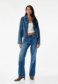 Chaqueta de mezclilla azul con un diseño de cuello, combinada con pantalones anchos de mezclilla azul y una camiseta corta blanca. Botas de tacón marrones completan el look.