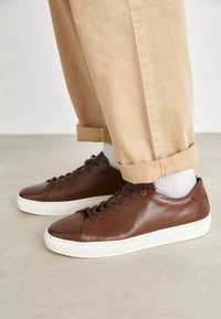 Sneakers in pelle marrone con suole in gomma bianche, caratterizzati da punte rotonde, lacci anteriori e superficie texturizzata. Indossati con pantaloni beige arrotolati.