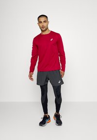 Červené dlouhé rukávy sportovní tričko, šedé šortky, černé legíny s vzorem, černé běžecké boty s oranžovými detaily, ve stoje.