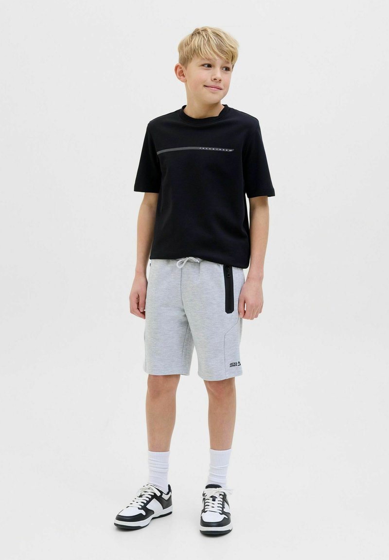 Maglietta nera a maniche corte con una striscia grigia orizzontale sul petto, abbinata a pantaloni corti grigi con tasche con zipper e dettagli neri. Sneakers bianco e nere.