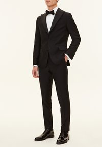 Svart smoking med satinrevär, vit plisserad skjorta och svart fluga. Glansiga svarta läderskor fulländar outfiten.