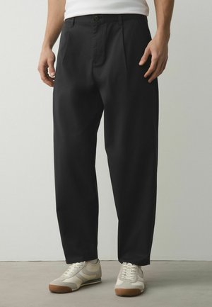 Mann trägt lockere schwarze Hose mit Falten, weiße Sneakers mit Gummisohlen und ein weißes eingestecktes Hemd, steht auf grauem Boden.
