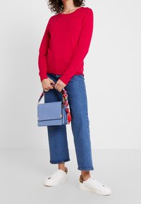 Rood top met lange mouwen, blauwe wijde jeans, witte sneakers met luipaardprintaccent. Houdt een blauwe tas vast met een kleurrijke sjaal.