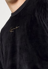 Černá sametová košile s dlouhými rukávy a kulatým výstřihem. Na levé straně hrudi se nachází vyvýšené zlaté výšivkové logo. Měkká textura.