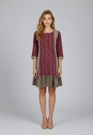 Mujer de pie con un vestido hasta la rodilla de color granate y beige con estampado, mangas tres cuartos y tacones beige sobre fondo gris.