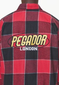 Czerwona i czarna kraciasta koszula z kołnierzem, z dużym haftowanym logo "PEGADOR LONDON" w żółtym i białym kolorze na plecach.