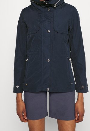 Marineblauwe capuchonjas met drukknoopsluiting aan de voorkant, borst- en zijzakken, gedragen met donkergrijze korte broek.