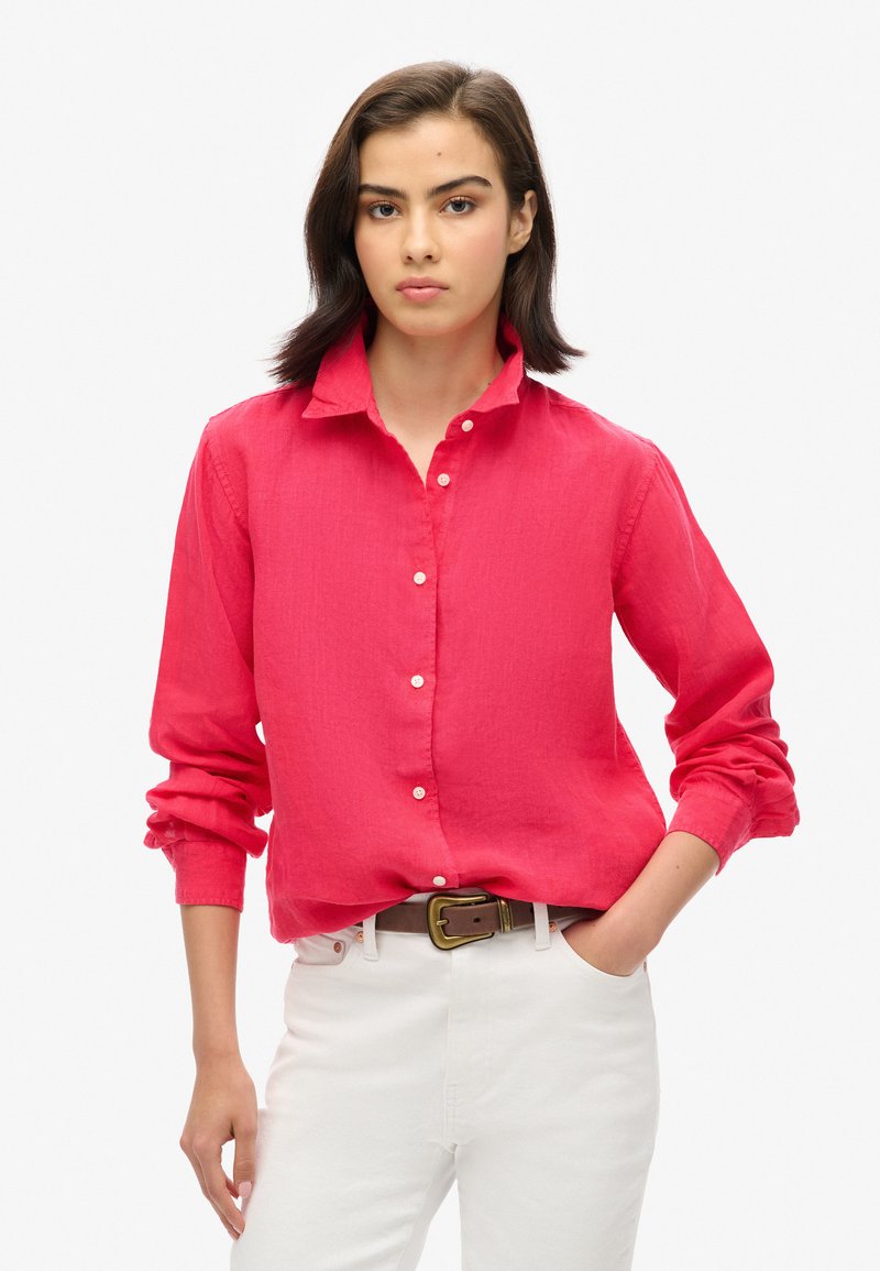 Roze overhemddetail van lichtgewicht stof, met een kraag en opgerolde mouwen, gecombineerd met een witte broek en een bruine riem.