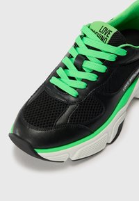 Zwarte atleet sneakers met groene veters en accenten. Bevat mesh en leren materialen. Getextureerde zool voor grip en ondersteuning. Label zichtbaar.