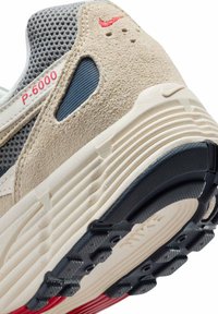Beige sneaker with gray mesh upper, suede accents, and red logo detail. Textured outsole features ridges and grip patterns. Model designation visible.