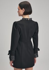 Robe noire avec une silhouette ajustée, des manches bouffantes et un col à pois. Des poignets froncés avec des accents à nouer complètent le design.