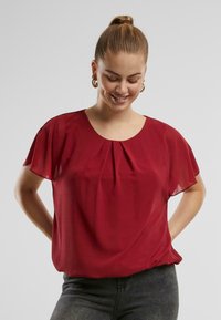 Rode blouse met korte mouwen en ronde hals, voorzien van plooidetails. Gemaakt van lichte stof met een zachte textuur. Draagt bij donkerblauwe jeans.