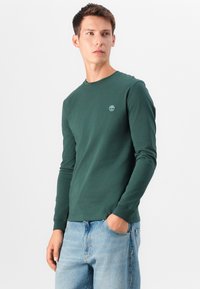 Langærmet grøn bomulds T-shirt med rund hals og lille logo på brystet, parret med lyseblå denim jeans.