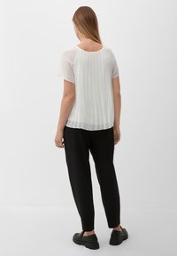 Blouse blanche plissée à manches courtes et col rond, portée avec un pantalon noir fuselé et des chaussures noires, vue de dos.