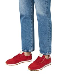 Scarpe da ginnastica rosse con un mix di materiali lisci e scamosciati, suole in gomma piatta e una linguetta con marchio di colore chiaro, abbinate a jeans blu.